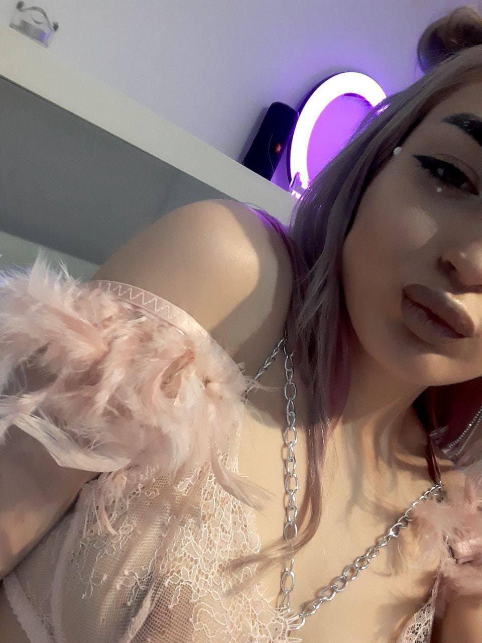 Close-up selfie with light pink hair, sheer lace top with feather trim, and silver chain, lit by a purple ring light.
