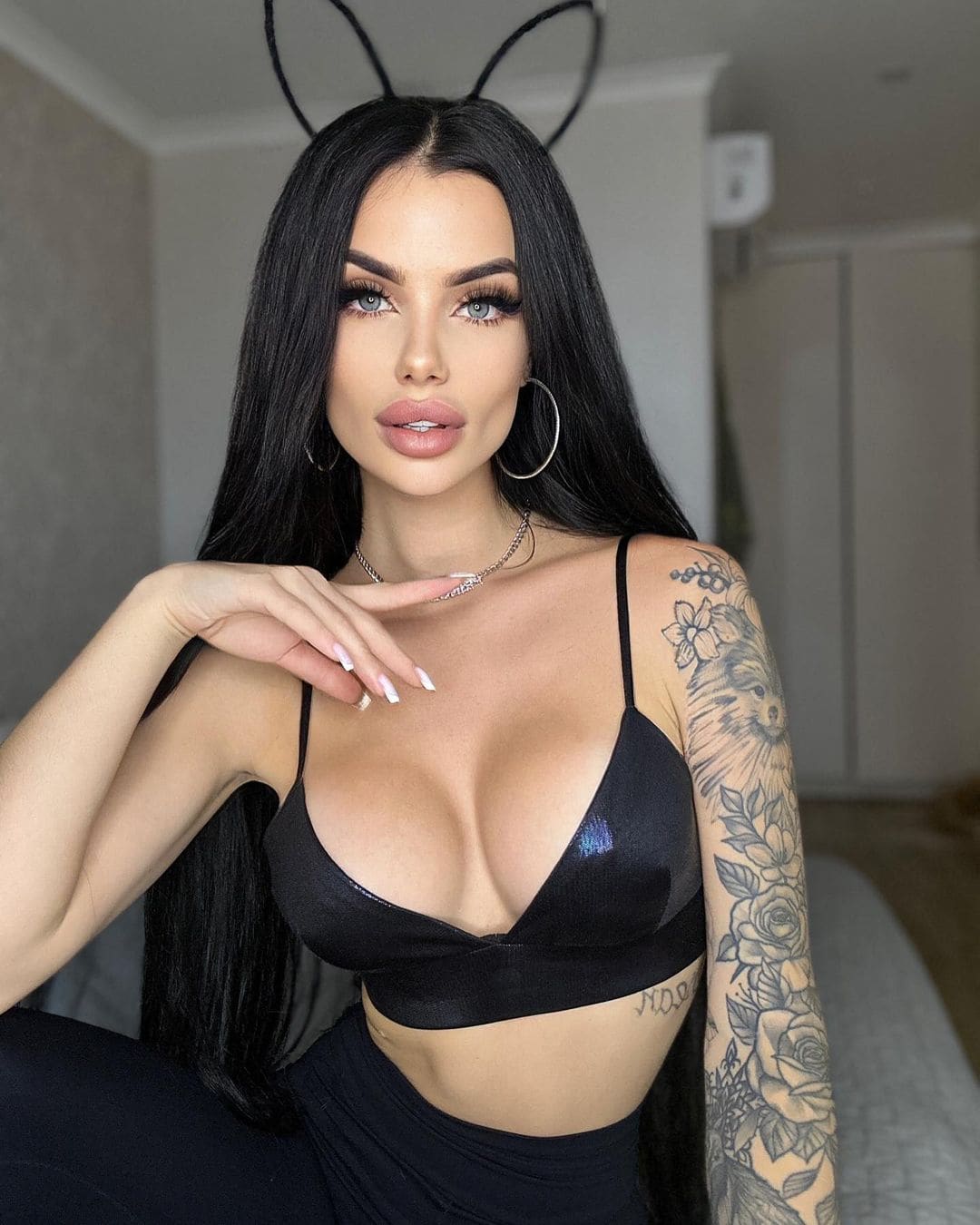 Sofia indoors in a black bralette and bottoms, hand near chest, floral tattoo and hoop earrings visible.