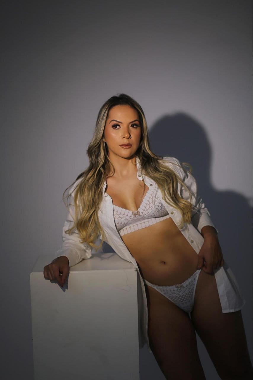 A blonde woman in white lace lingerie and an open white shirt leans on a pedestal, looking at the camera.