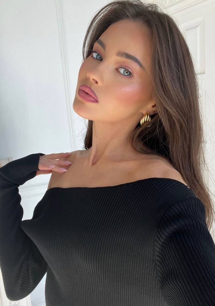 Nicole wears a black off-the-shoulder ribbed sweater and gold hoops, posing with a hand on her shoulder against a white paneled wall.