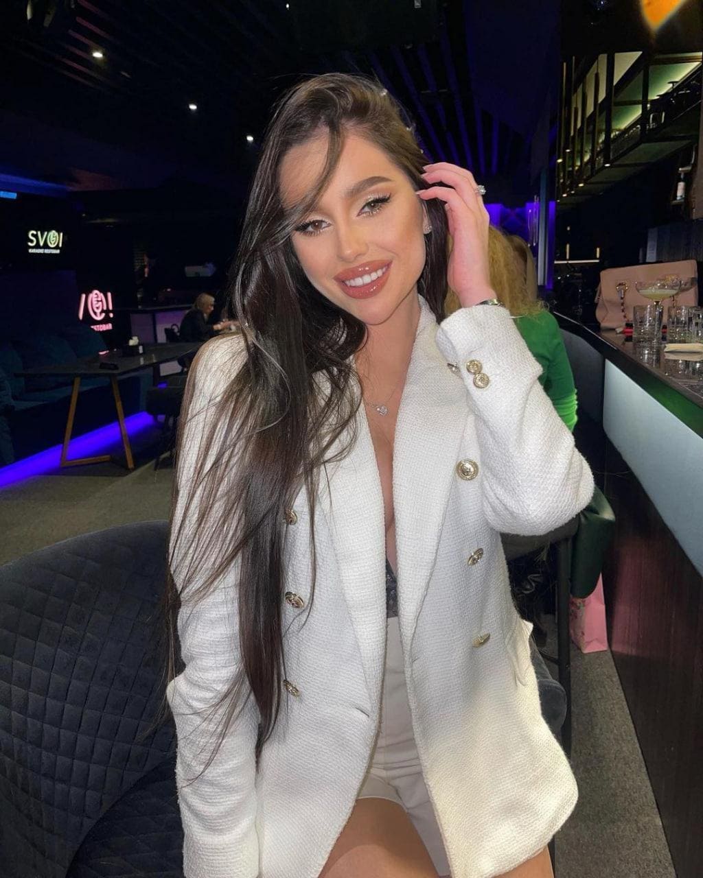 Kylie sits in a dim lounge wearing a white textured blazer with gold buttons, smiling with her hand raised to her hair.