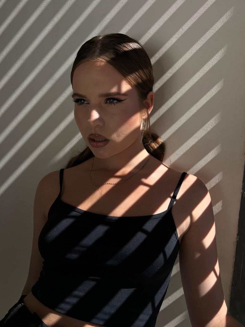 A woman with dark hair in a black crop top and necklace stands against a white wall, shadows from blinds crossing her face.
