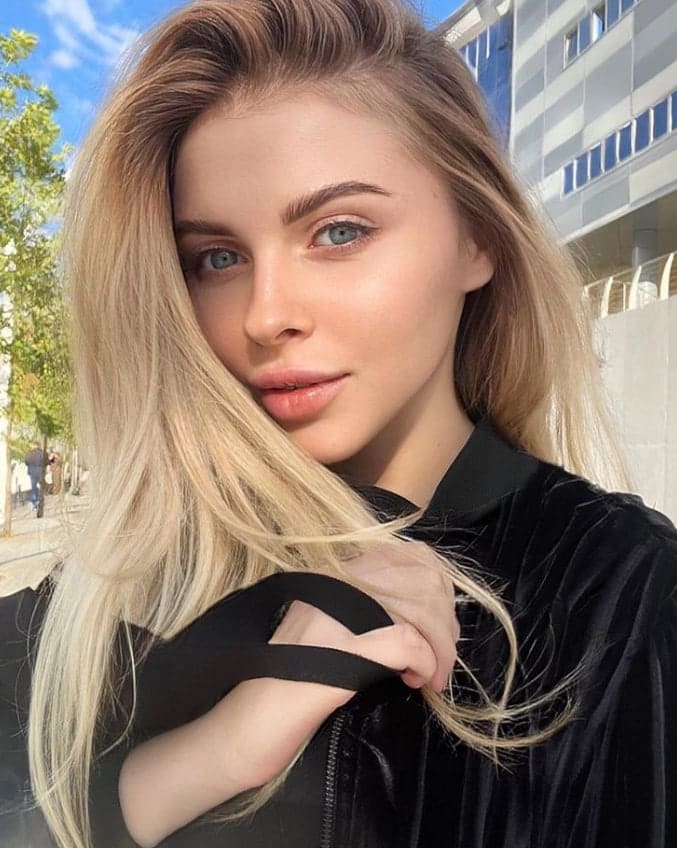 Eliza in a black velvet top with shoulder cutout, standing outdoors before a modern building and trees under a blue sky.