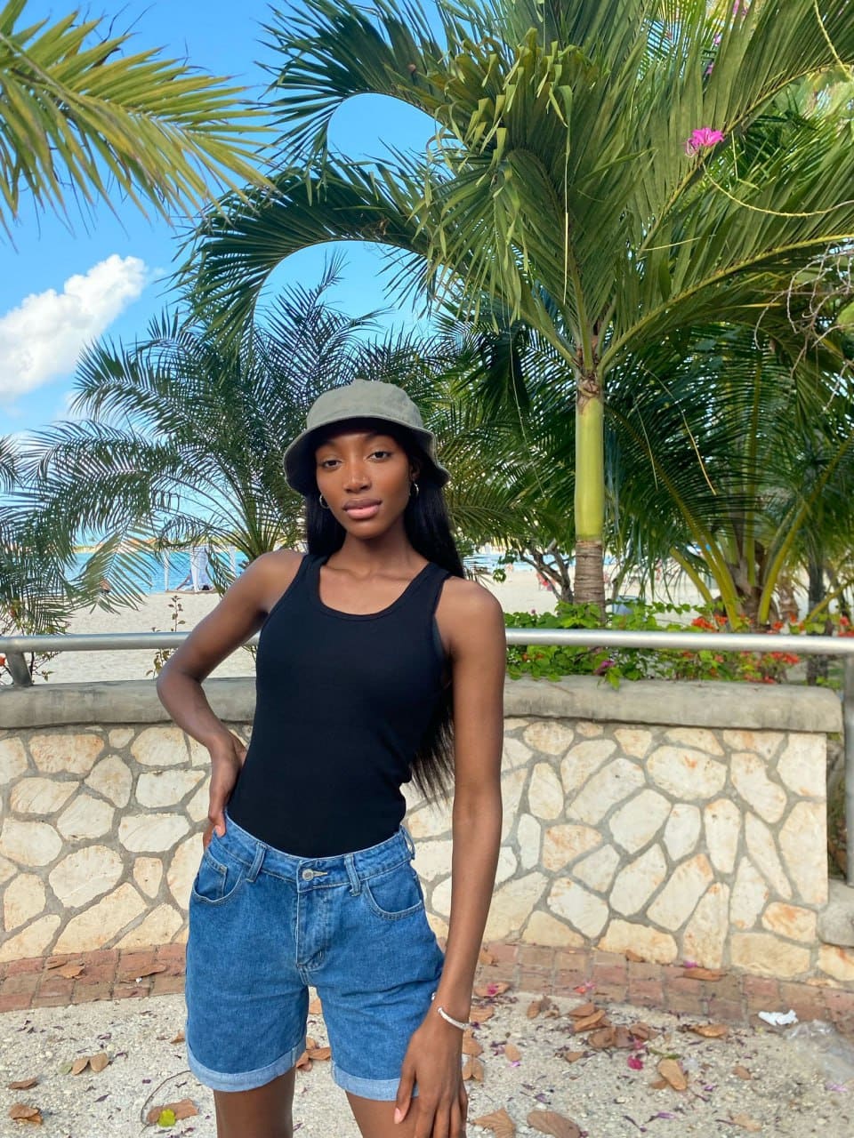 A woman in a grey bucket hat, black tank, and denim shorts poses outdoors near palm trees and stone.