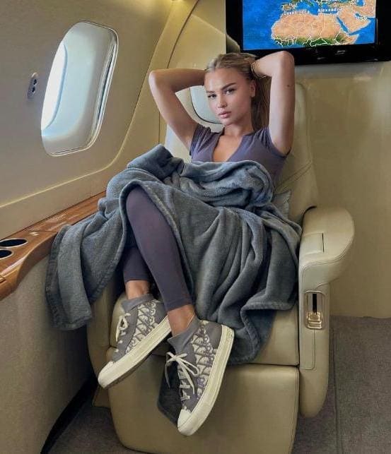 A woman sits in a private jet seat wearing a grey top, leggings, and sneakers, with a blanket on her legs and hands behind her head.