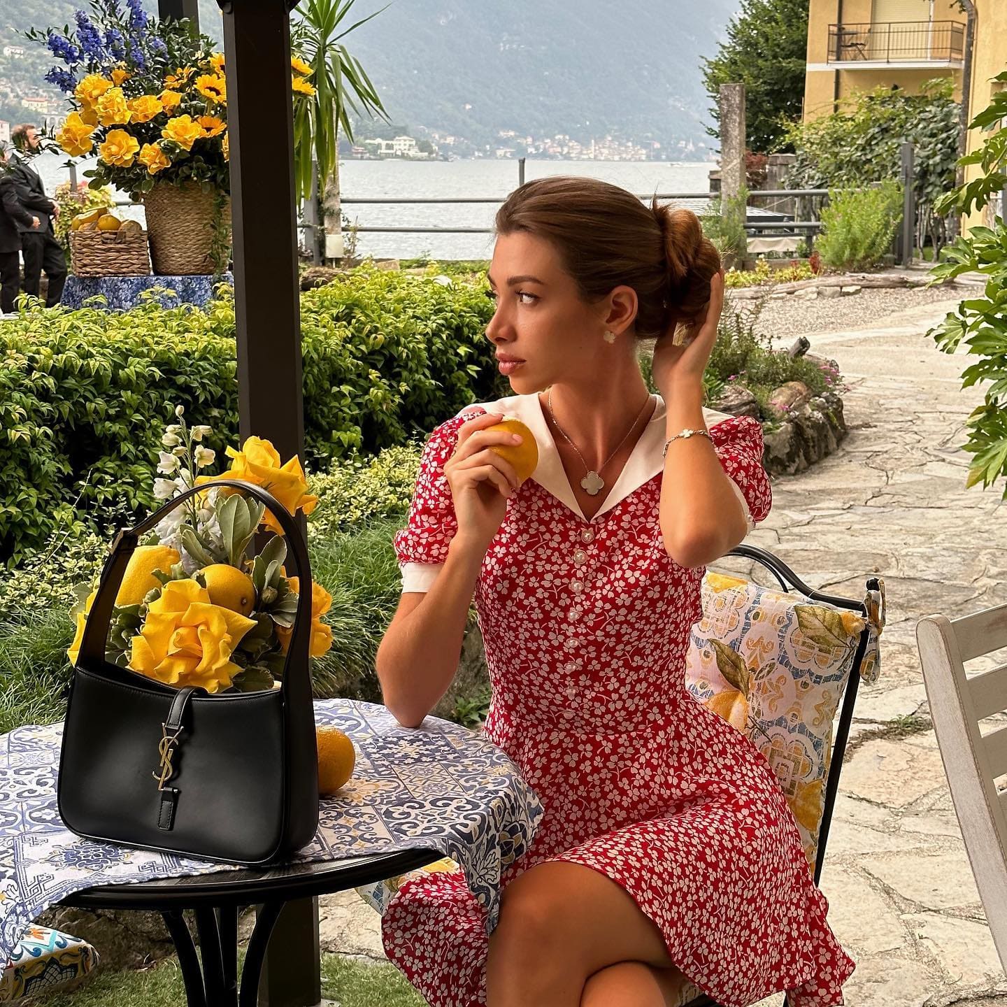 A woman in a red floral dress sits at an outdoor table holding a lemon, with a black handbag and yellow flowers nearby.
