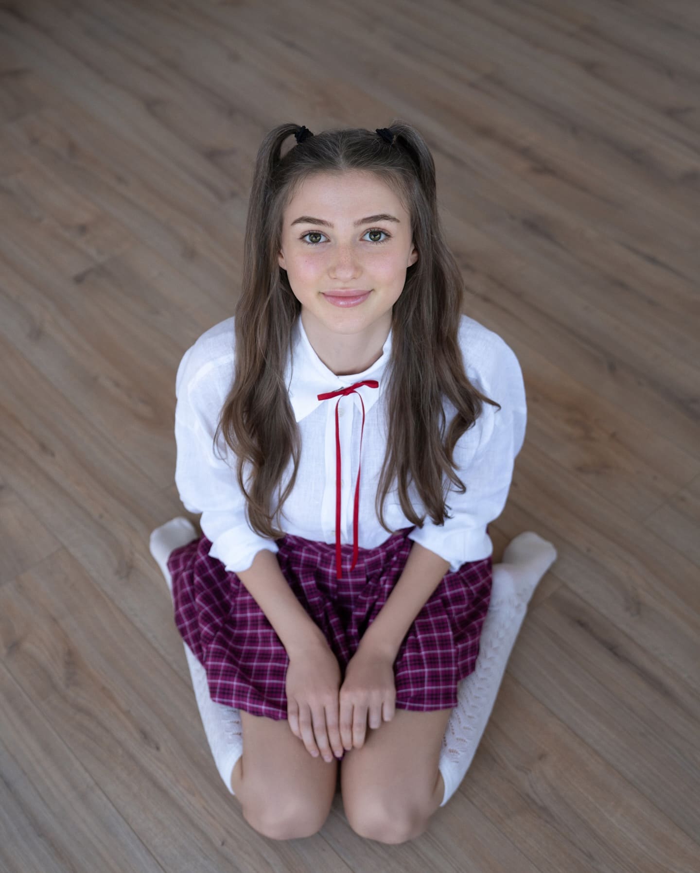 Sitting on a wooden floor in pigtails, wearing a white blouse with red ribbon and purple plaid skirt, looking up.