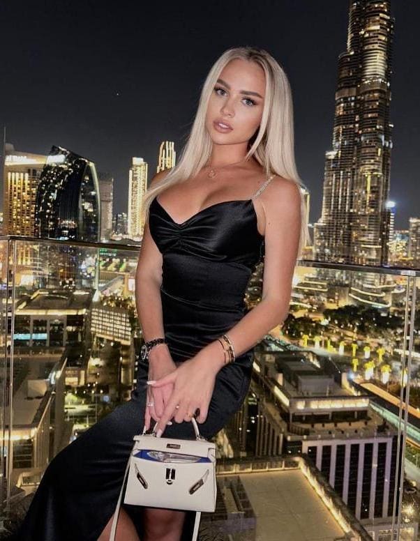 A woman in a black satin slip dress sits on a rooftop ledge at night, holding a white handbag, with a glowing city skyline visible in the background.