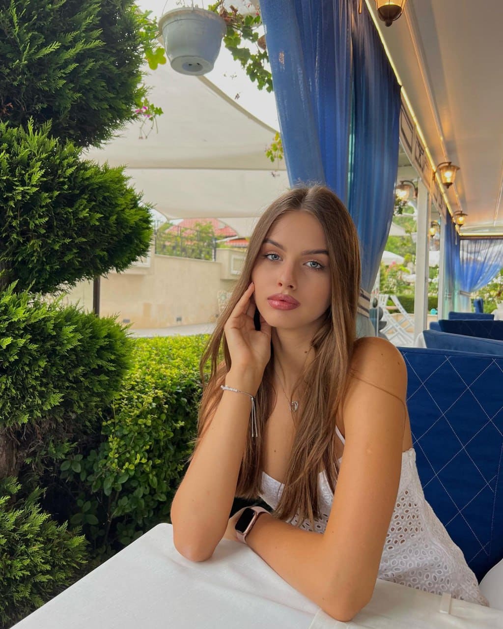 Amelia sits at a white cafe table in a white lace top, hand on cheek, with green shrubbery and blue curtains behind her.