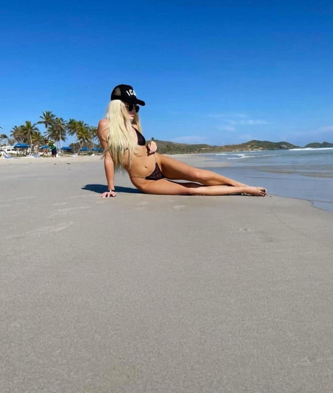 A woman in a black baseball cap, sunglasses, and black bikini sits on a sandy beach with legs extended.