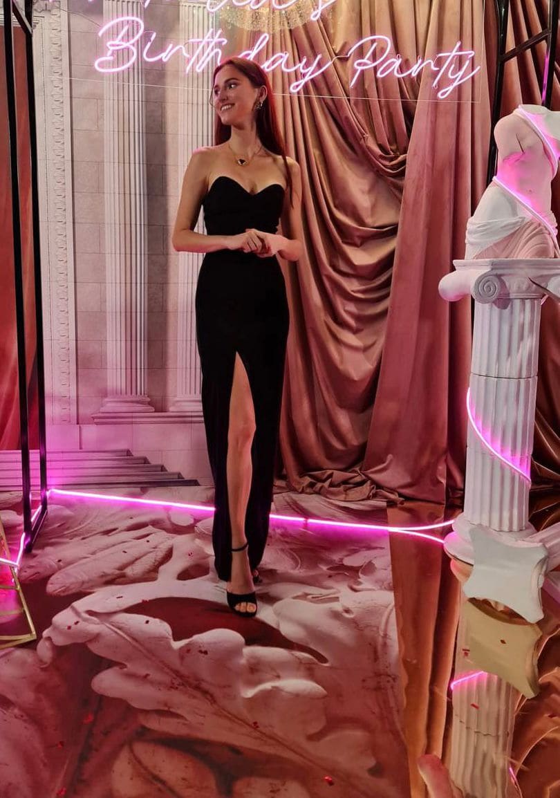 Standing before a pink-lit backdrop with a neon sign, she wears a black strapless gown with a high front slit and heeled sandals.