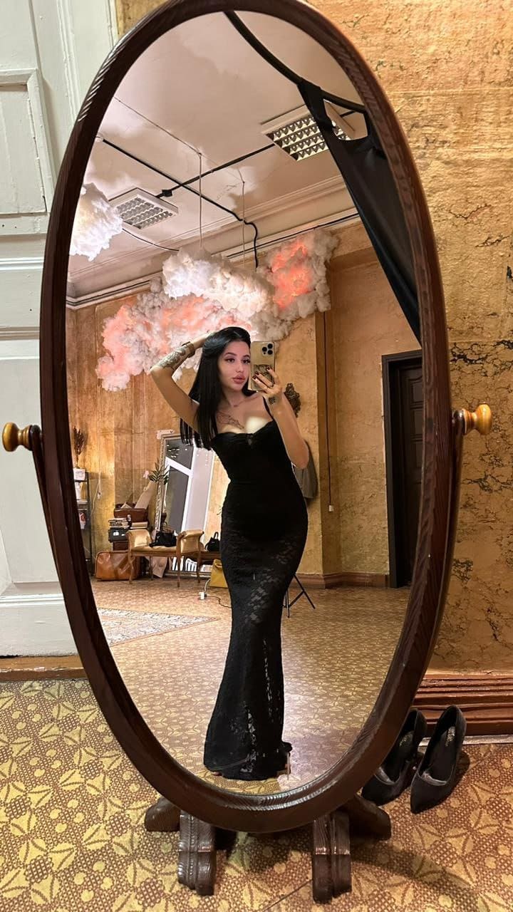 A woman in a sleeveless black lace floor-length dress stands before an oval mirror, holding a phone and touching her hair in a room with patterned tiles.