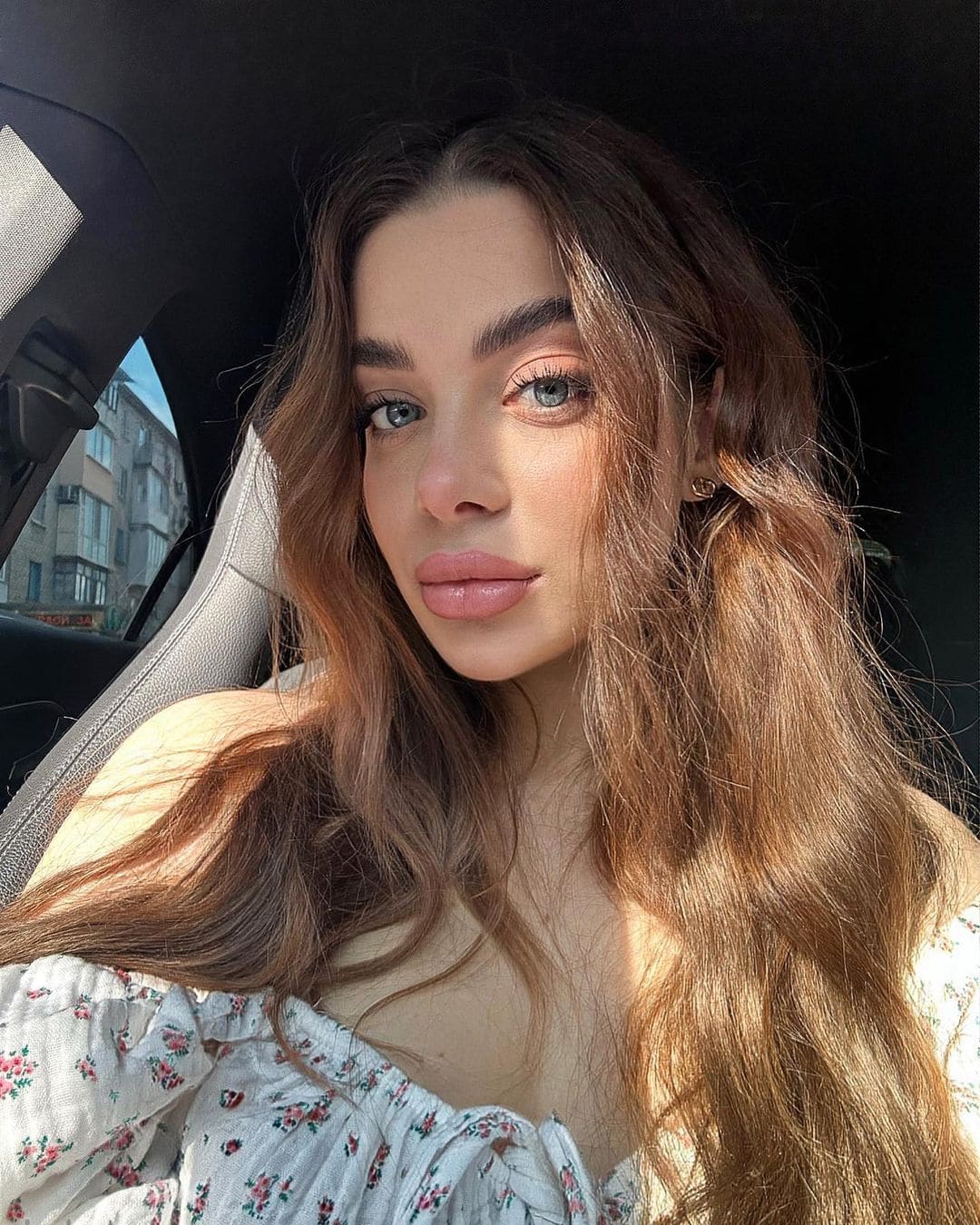 A woman sits in a car passenger seat wearing a white off-the-shoulder top with a small floral print, facing the camera with buildings visible behind her.