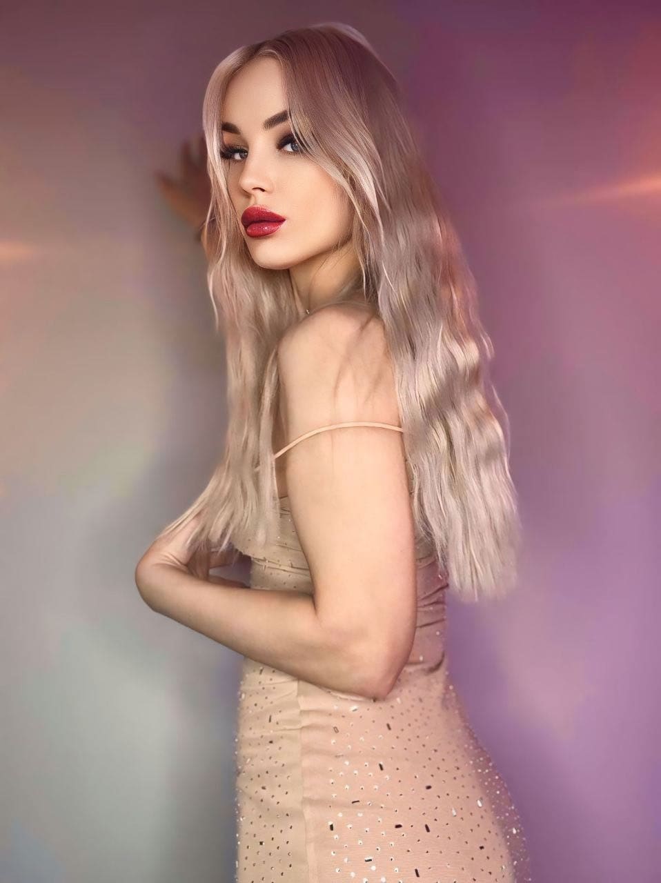 A woman in a form-fitting beige dress with sequin details stands in profile with arms crossed against a soft purple and pink gradient backdrop.