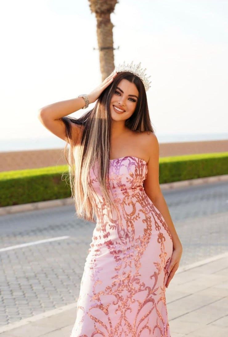 Nina stands outdoors near a palm tree, wearing a strapless pink and gold gown and tiara, with one hand raised to her head.