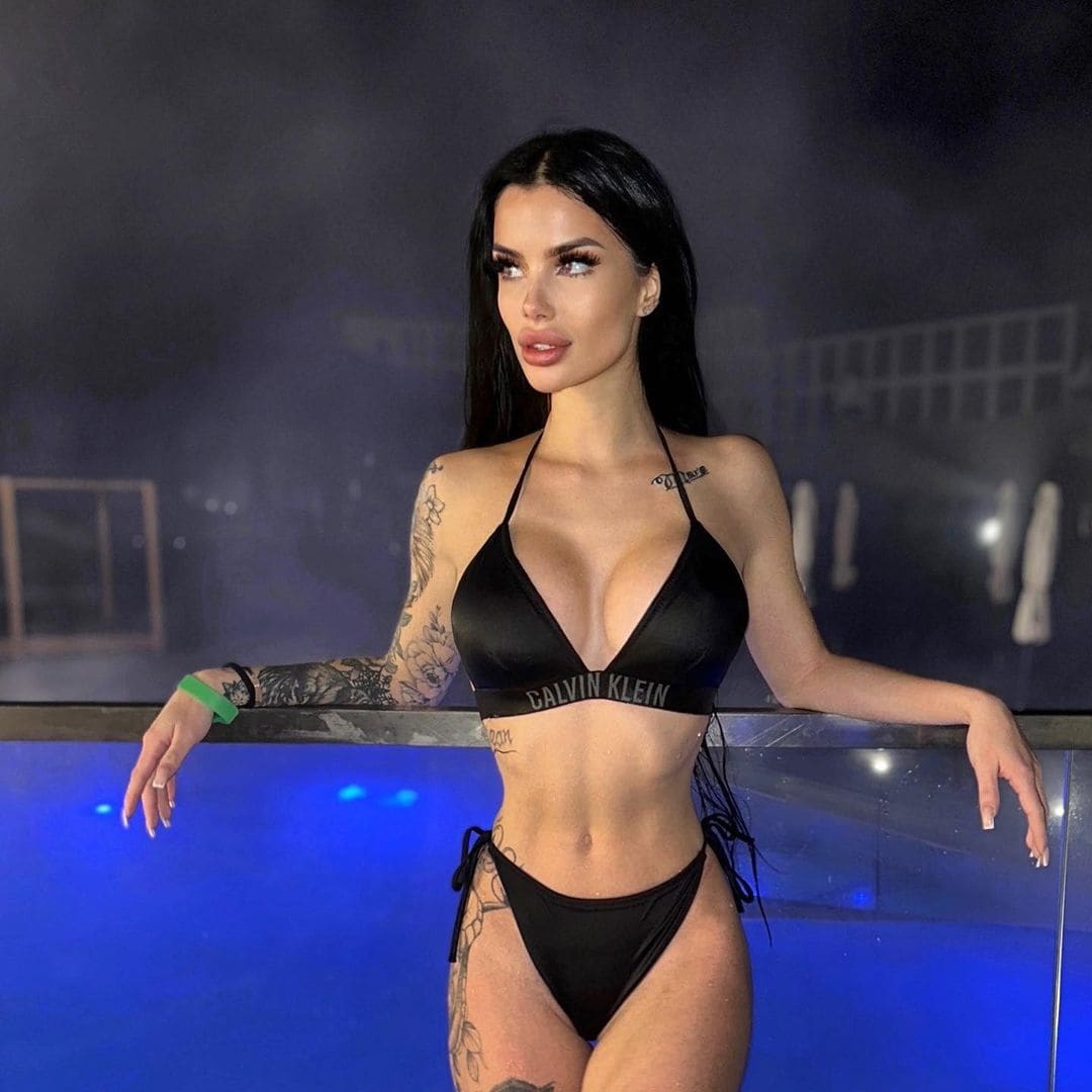 Woman in black two-piece bikini leaning on glass railing above blue-lit pool at night, tattoos visible.