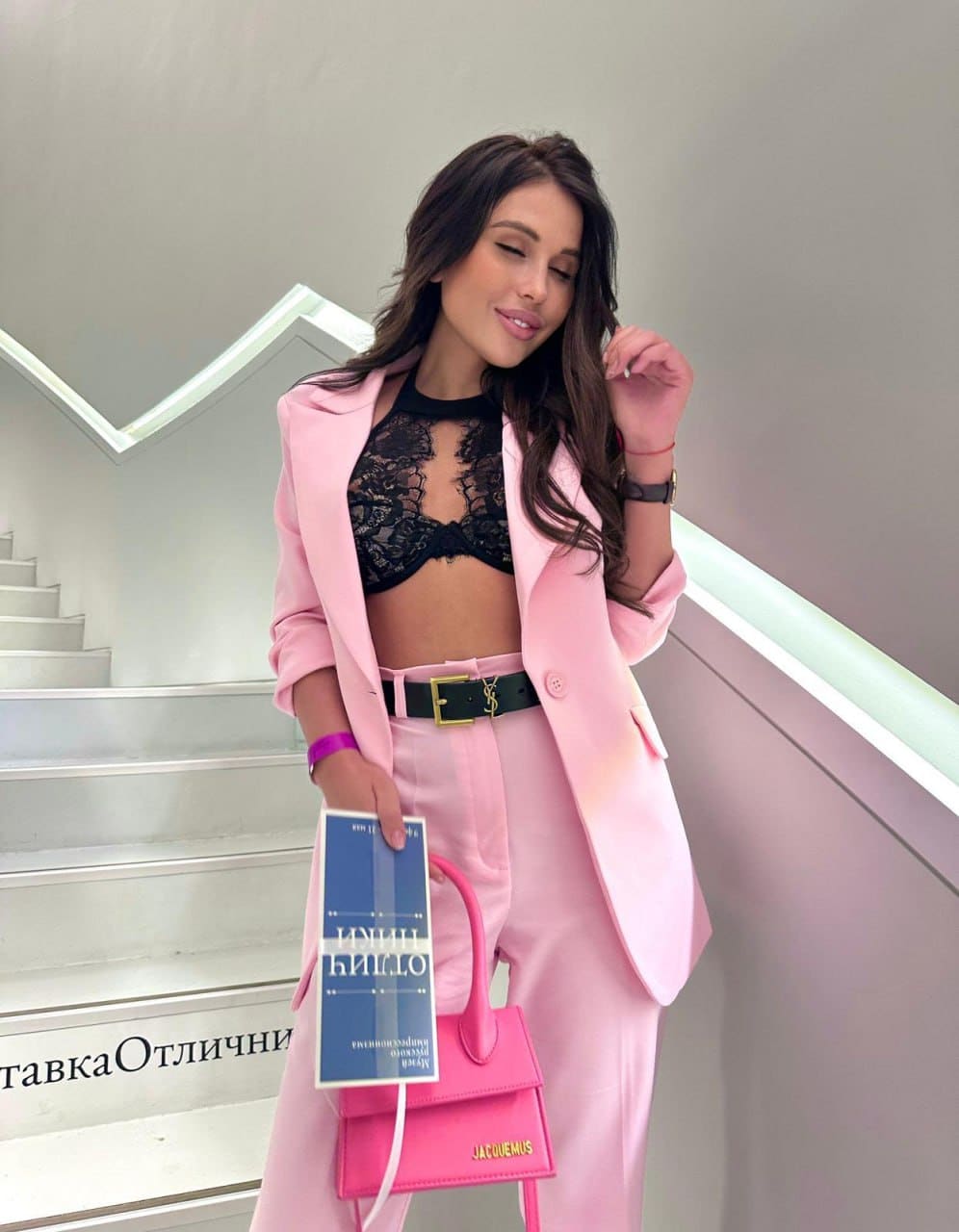 Anny stands on a white staircase in a pink suit and black lace top, holding a handbag and book while looking down.