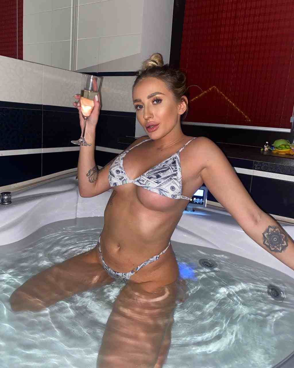 A blonde woman in a dollar-print bikini stands in a jacuzzi holding a champagne flute against tiled walls.