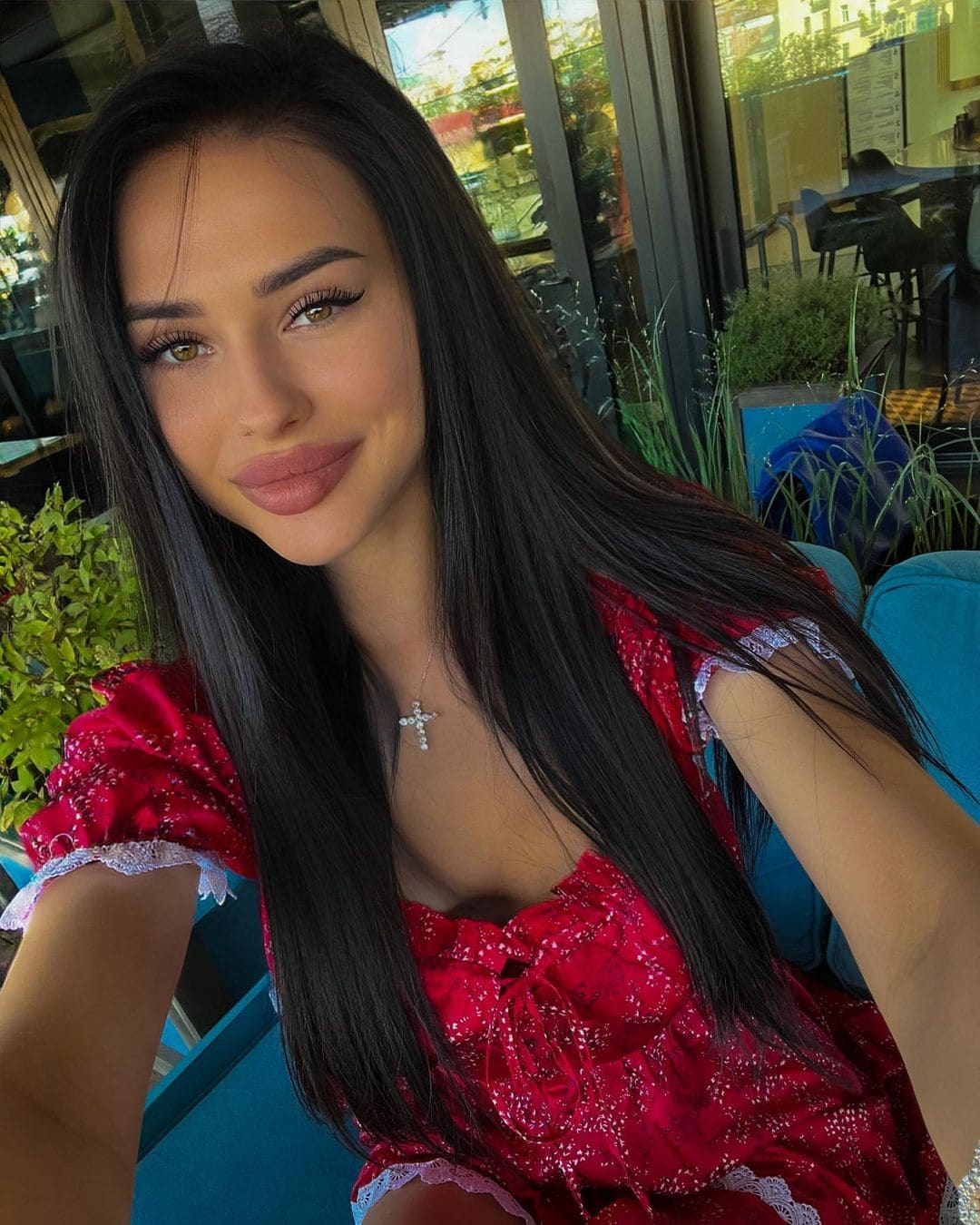 Betsy takes a selfie in a red sequined dress with lace sleeves, seated on a blue chair in a sunlit room with plants.