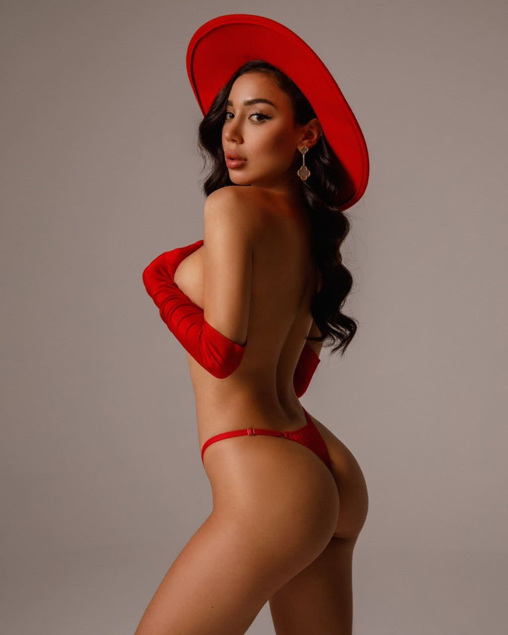 A woman stands in profile against a gray backdrop wearing a red wide-brimmed hat, red off-shoulder top, and red bikini bottoms, back partially turned.