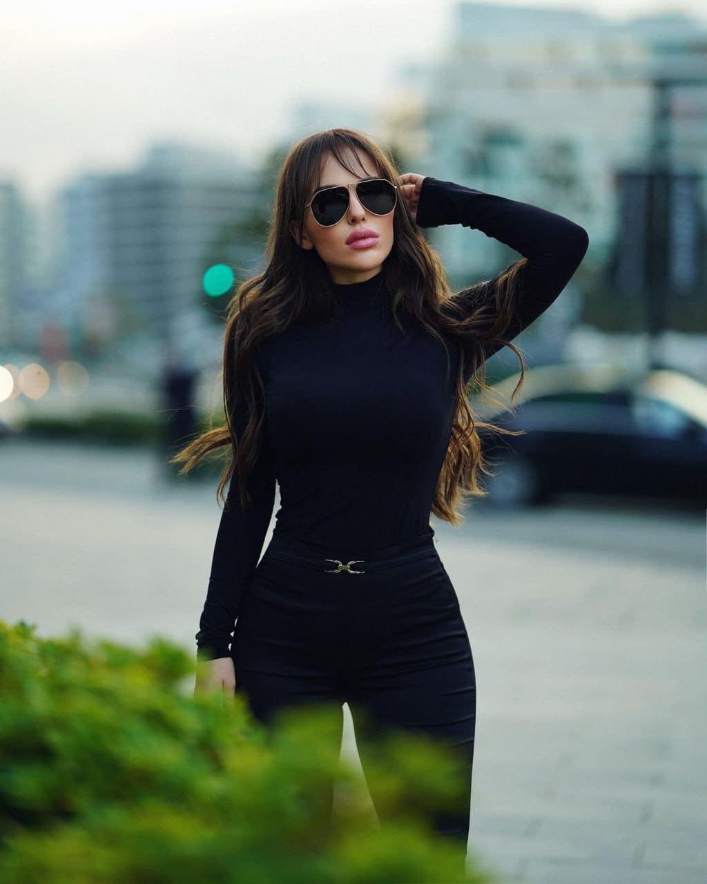 A woman with bangs wears a black turtleneck and pants with a metal buckle, one hand raised to her head, standing outdoors with blurred city streets and greenery.