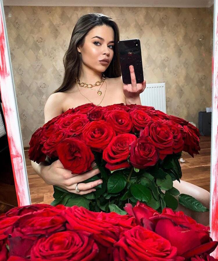 A mirror selfie with a large bouquet of red roses covering the torso, wearing gold jewelry in a room with patterned wallpaper.
