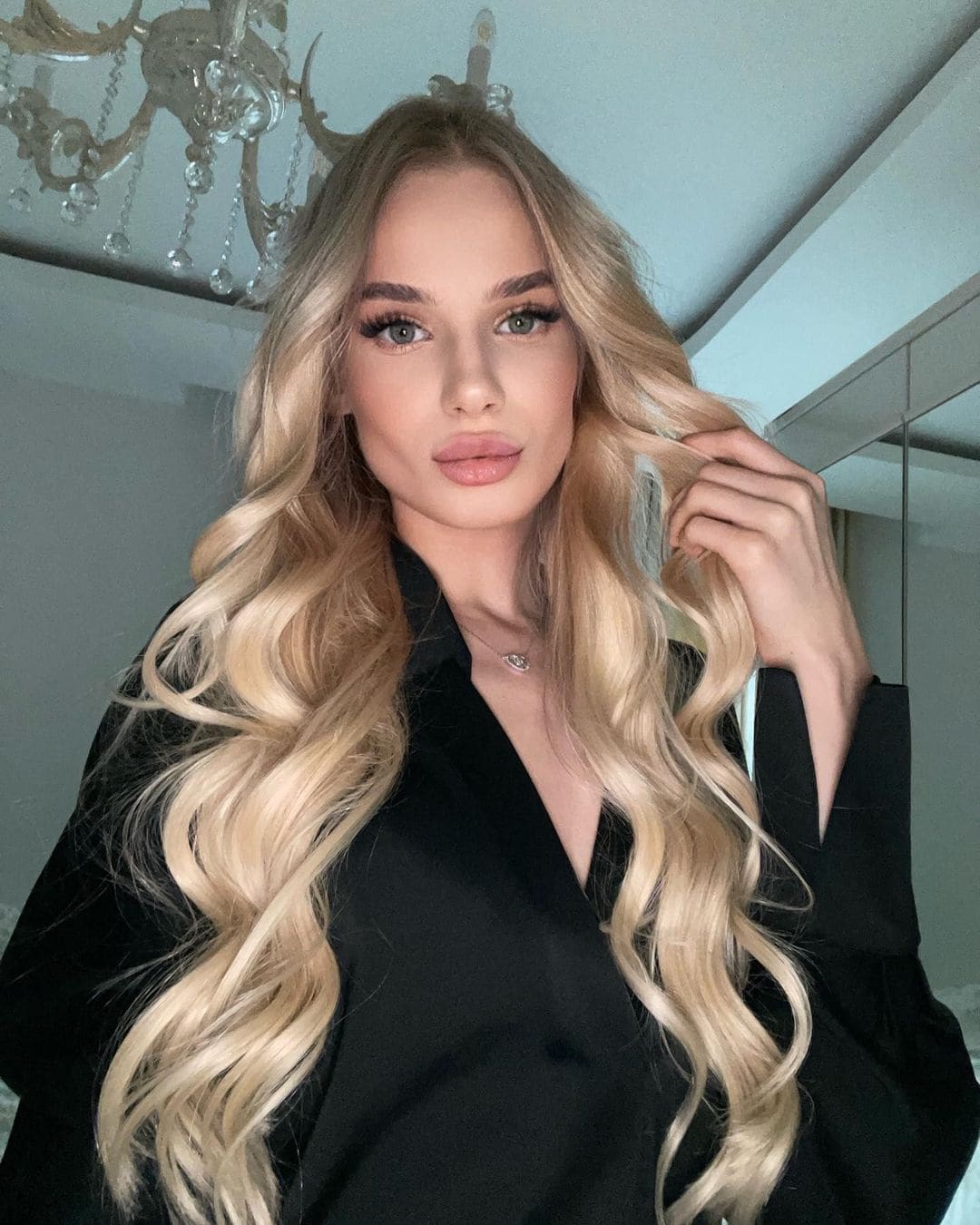 Rosalie stands indoors in front of a chandelier, wearing a black deep V-neck shirt and touching her wavy blonde hair.