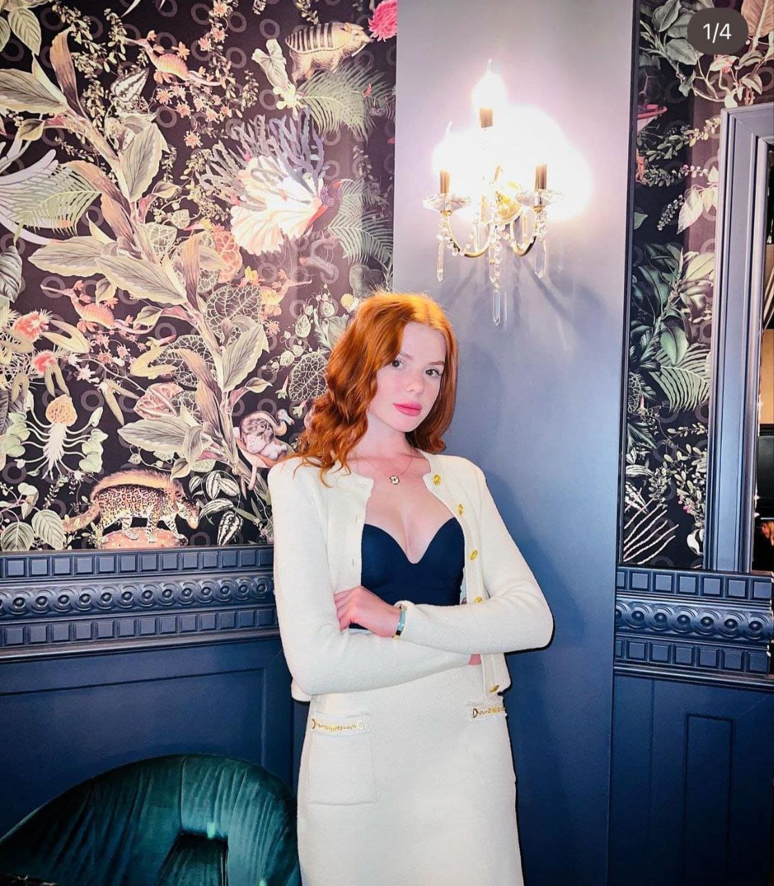 A red-haired woman stands with arms crossed in a white jacket and skirt, against dark floral wallpaper and a lit wall sconce.