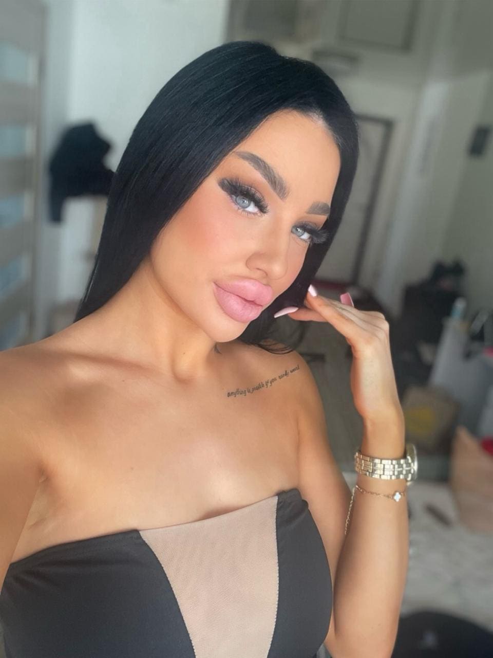 Agnes with long black hair and heavy makeup poses indoors in a strapless black and beige top, hand near chin, wearing gold jewelry.