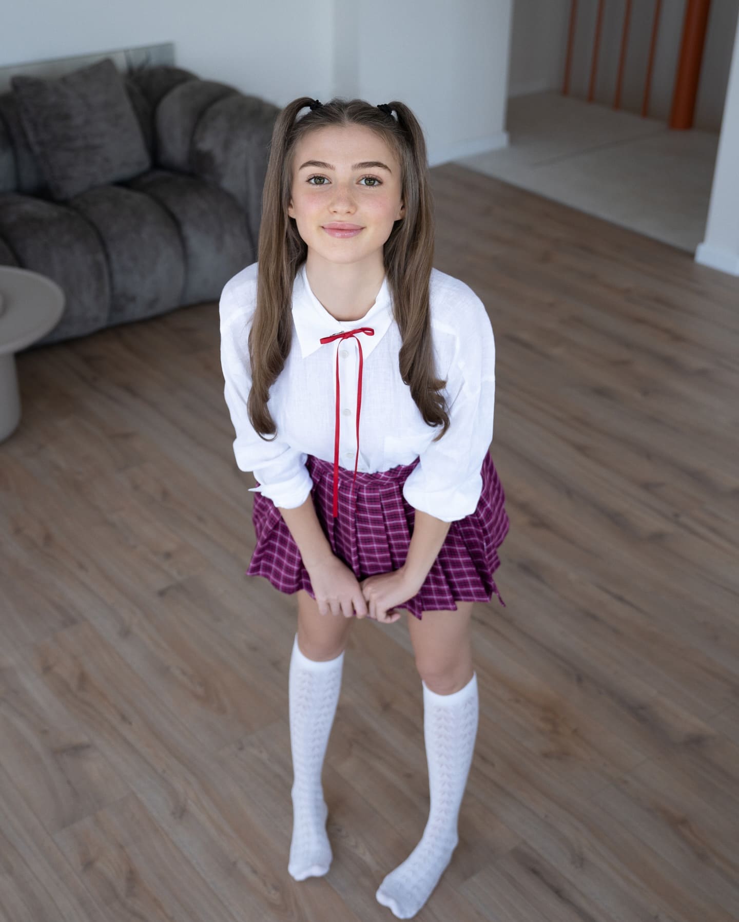 Standing on a wooden floor in pigtails, wearing a white shirt with red ribbon, purple plaid skirt, and knee-high socks.