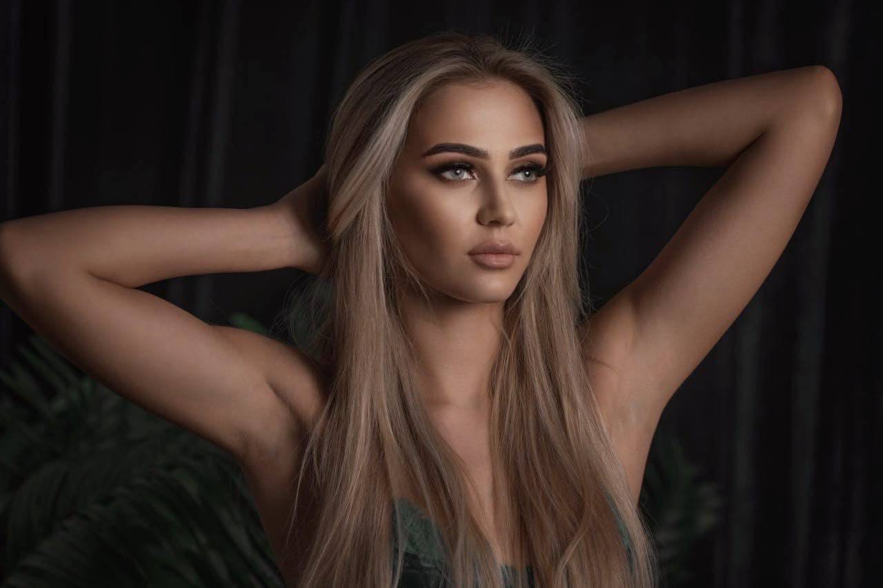 Solenne raises both arms behind her head, wearing a dark green strapless top against a dark background with blurred foliage.