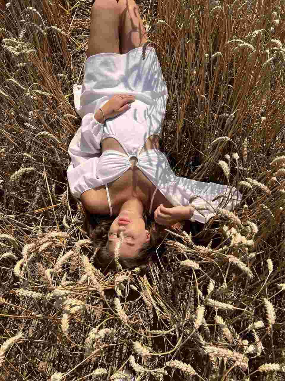 A woman lies in dry wheat wearing an open white long-sleeved top over a white bra, legs bent, one hand on her chest.