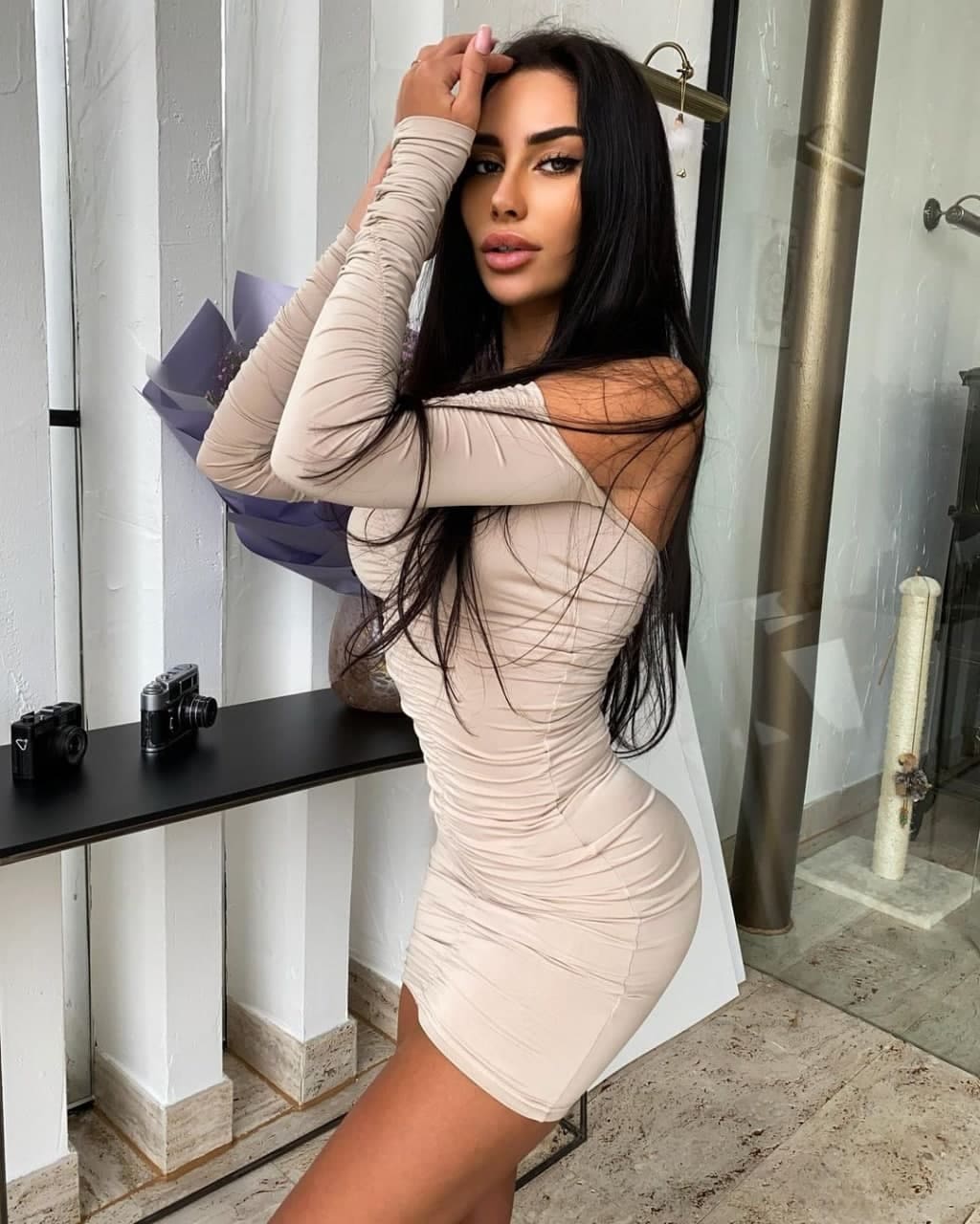 Edita stands indoors in a beige off-shoulder mini dress beside a black shelf holding cameras and a bouquet.