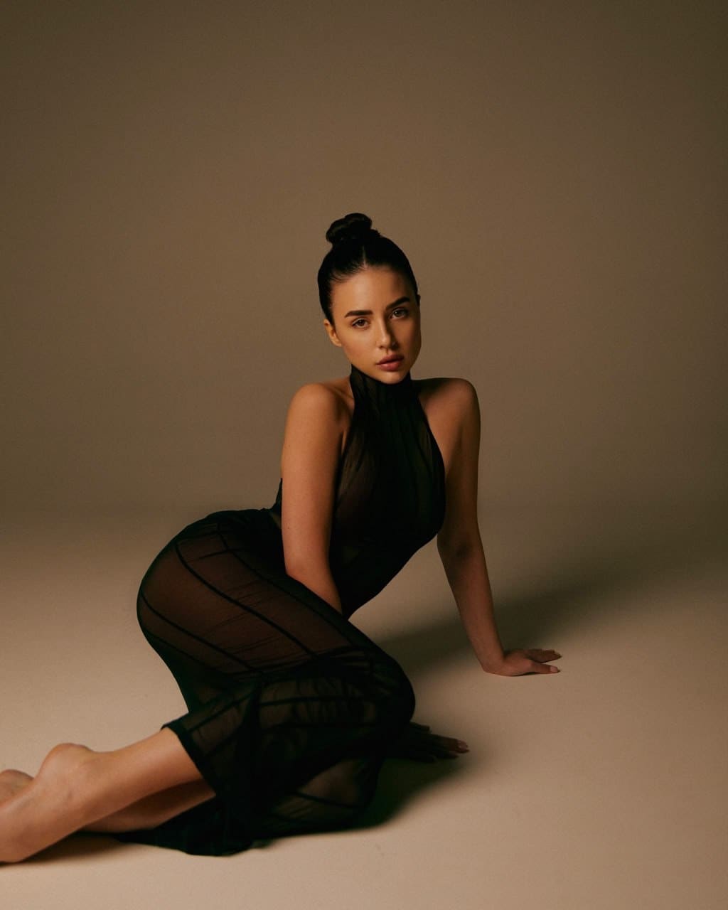 A woman kneels on a plain floor wearing a sheer black striped halter-neck dress, her right hand resting on the ground behind her.