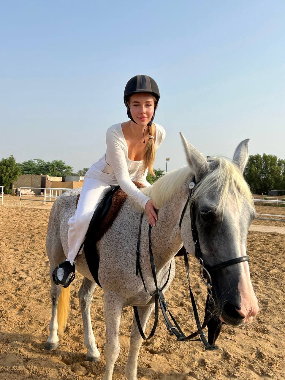 A woman with a blonde ponytail sits on a grey horse in an arena, wearing a black helmet and white riding gear.