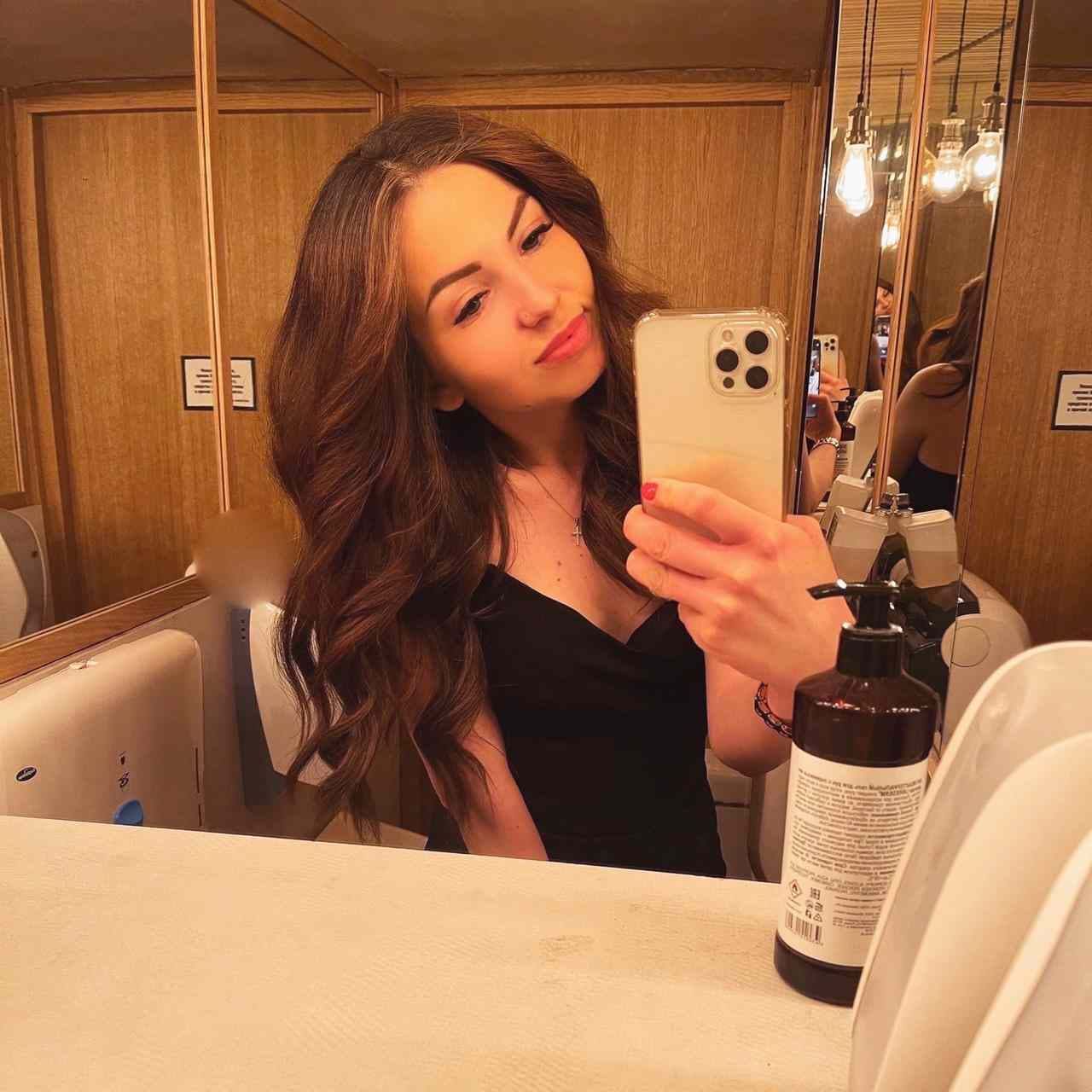 Mirror selfie in a wood-paneled room wearing a black sleeveless top, holding a white smartphone with a pump bottle nearby.