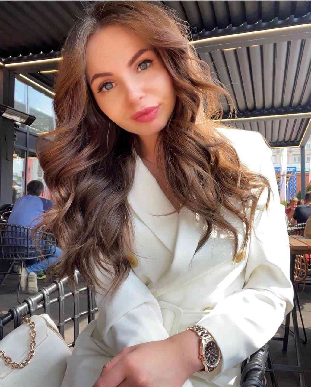 Ira sits at an outdoor cafe table in a white blazer, wearing a wristwatch with a white handbag beside her.