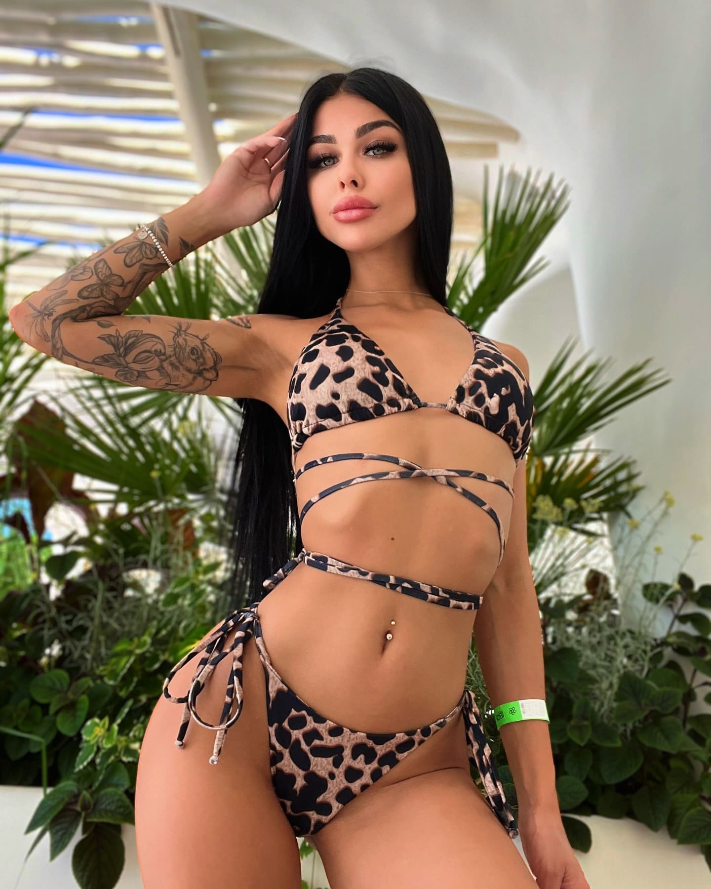 A woman with floral arm tattoos poses in a leopard-print strappy bikini, hand to head, standing before green plants under a slatted roof.