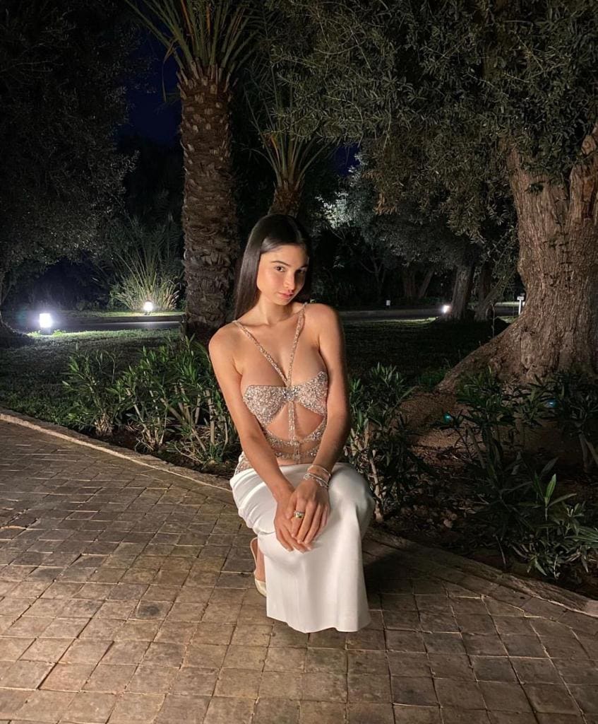 A woman kneels on a brick path at night in a sparkling cut-out top and long white skirt, with garden lights behind.
