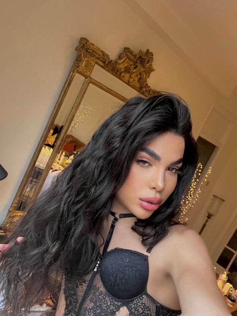 A woman poses in a black lace bodysuit with a halter neck strap, standing indoors in front of a large ornate gold-framed mirror.