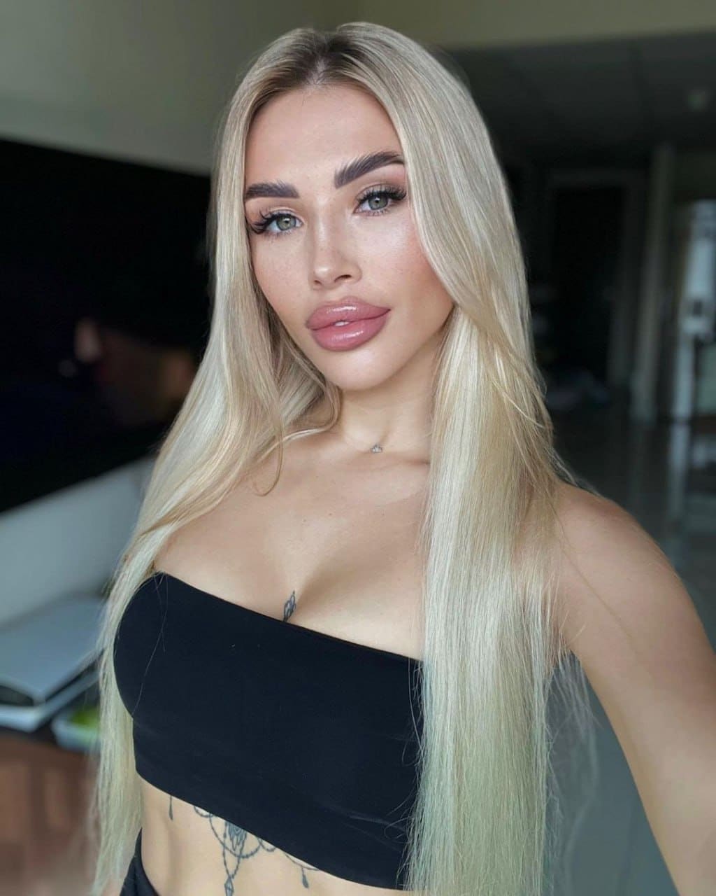 A blonde woman faces the camera in a black strapless tube top against a blurred indoor background.