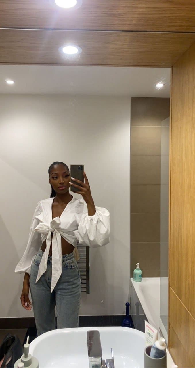A woman in a white tied shirt and blue jeans takes a mirror selfie in a bathroom with a sink.