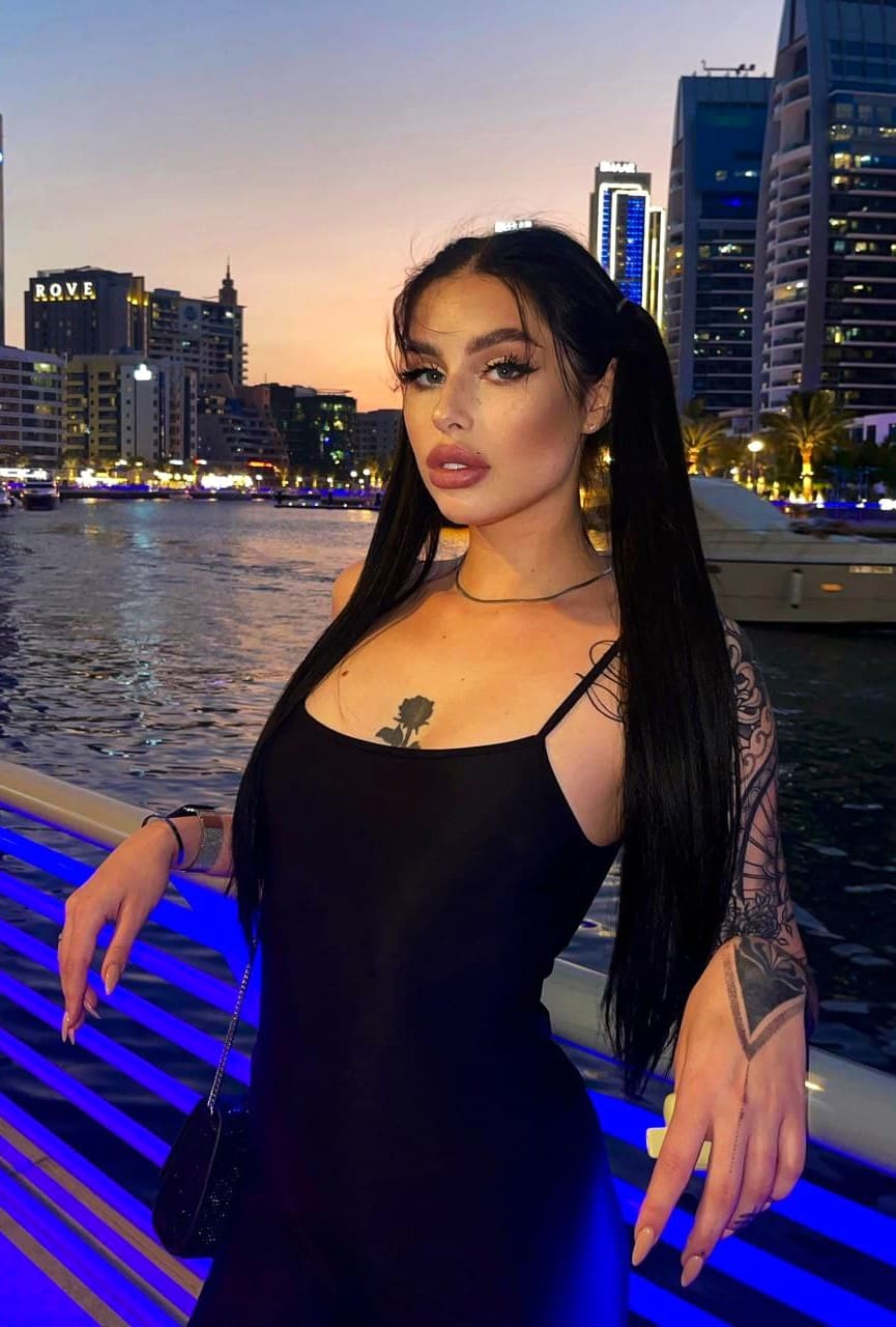 Standing on a waterfront promenade at dusk, she wears a black spaghetti-strap dress revealing a chest tattoo.