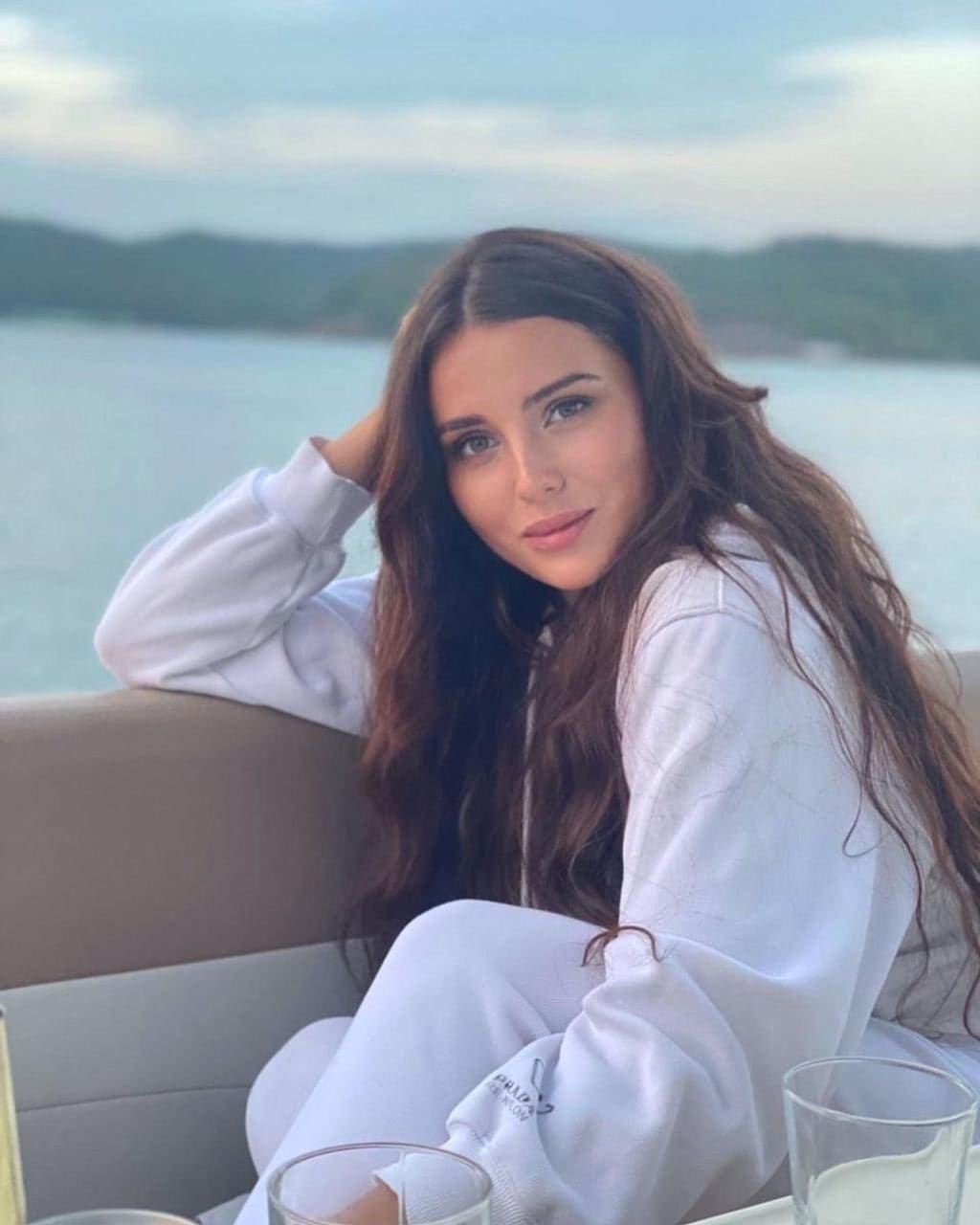 Albina sits on an outdoor bench in a white sweatsuit, hand behind her head, with water and hills in the background and glasses on a table.