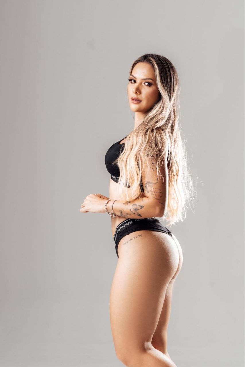 Profile shot of a blonde woman in a black bikini, hands clasped at waist, showing arm and hip tattoos against gray.