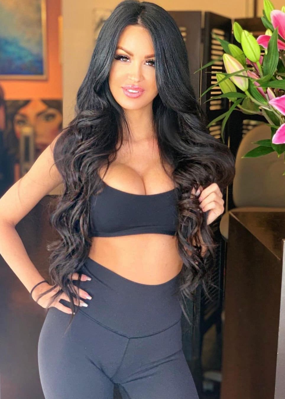 Jamie stands indoors in a black strapless crop top and leggings, hand on hip, with pink lilies and framed art behind her.