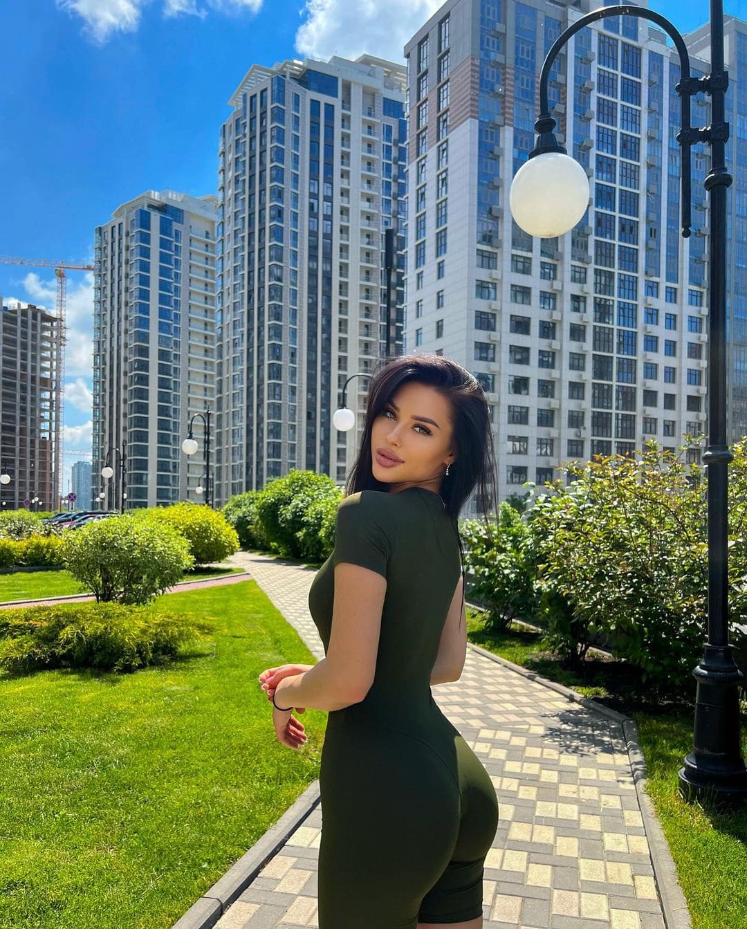 A woman stands in an urban park wearing an olive-green dress with an open back, with high-rise buildings in the background.