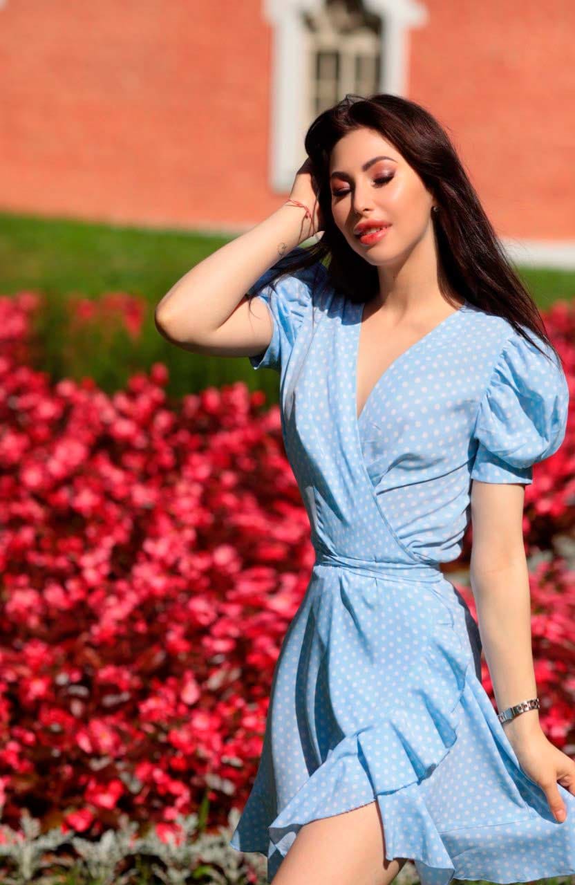 Amina stands outdoors in a light blue polka-dot dress with ruffled hem, one hand raised to her head, against a red brick building and pink flowers.
