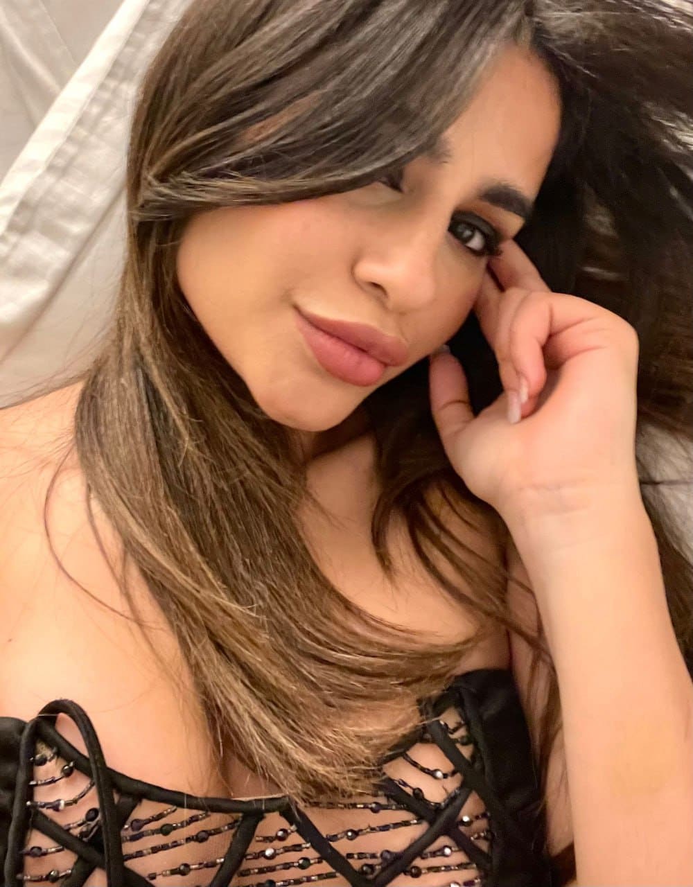 A woman rests her head on her hand in a black strappy top with beaded details, against a light-colored fabric background.