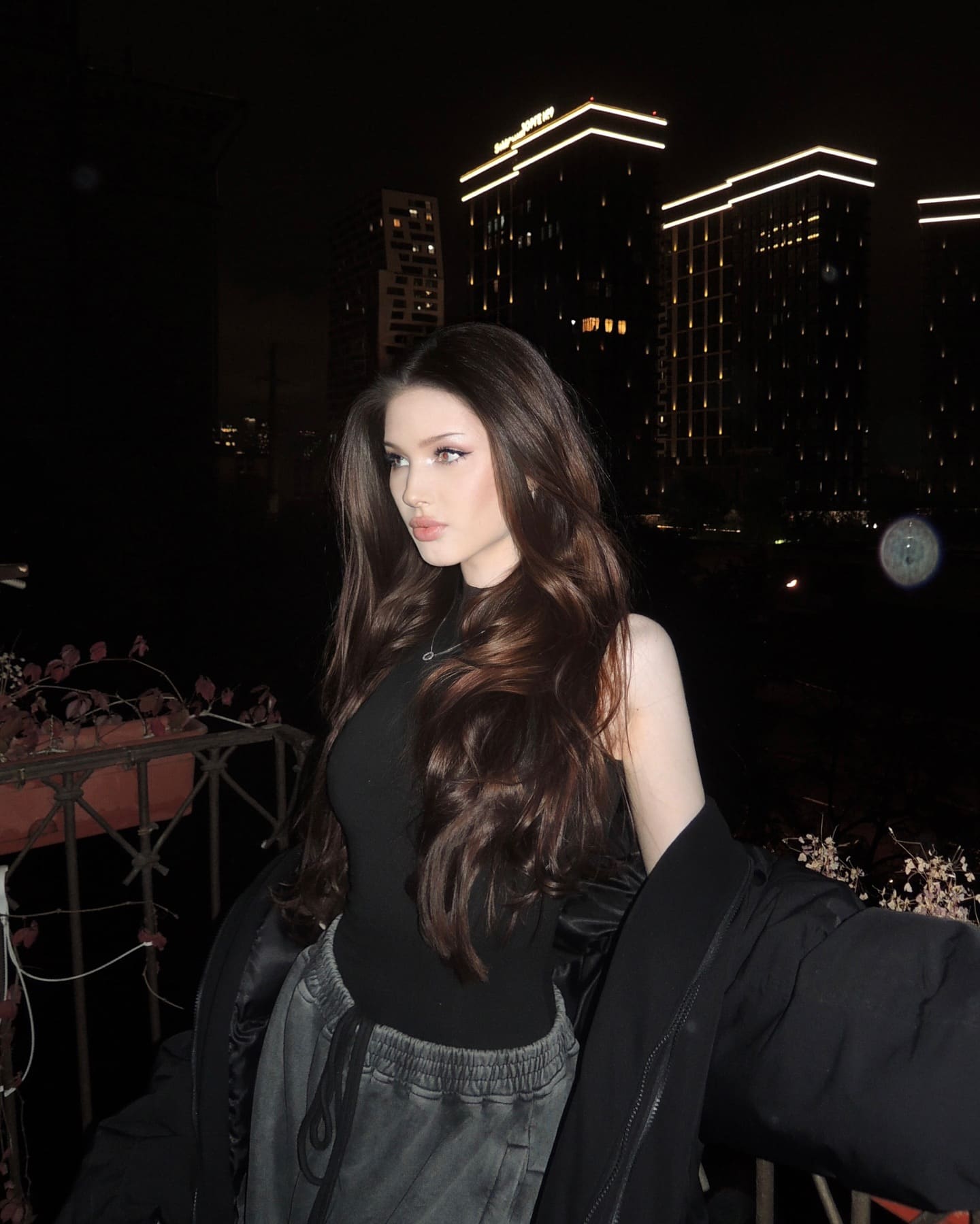 A woman in a black top, grey sweatpants, and draped jacket stands on a night balcony with city lights behind her.